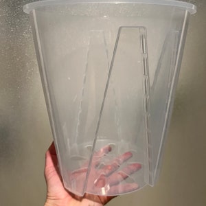 May include: A clear, transparent plastic plant pot with a wide top and a tapered design. The pot has a series of vertical supports on the interior and a drainage system at the bottom. The pot is held by a hand.