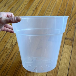 May include: A clear plastic plant pot with a wide, cylindrical shape. The pot has a slightly flared rim and a series of drainage holes at the bottom. The pot is held by a hand.