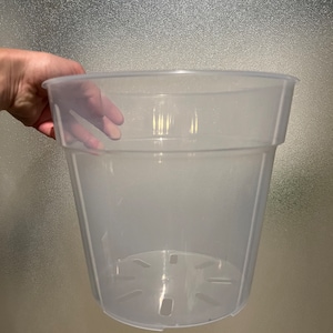 May include: A clear, transparent plastic plant pot with a wide rim and a tapered body. The pot has drainage holes in the bottom. The pot is held by a hand.