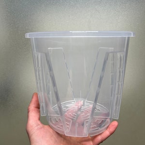 May include: A clear plastic plant pot with a square top and a patterned base. The pot has vertical supports and a slotted design for drainage. The pot is held in a hand.