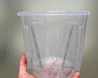 May include: A clear plastic plant pot with a square top and a patterned base. The pot has vertical supports and a slotted design for drainage. The pot is held in a hand.