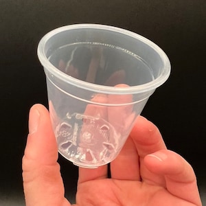 May include: A clear plastic plant pot with drainage holes at the bottom. The pot is small and is being held in a person's hand.