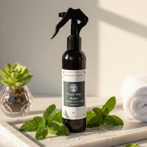Puede incluir: Un frasco rociador marrón oscuro con un gatillo negro, etiquetado como "From Tha Roots HYDRATION MIST Money Rush 8oz." Hojas de menta fresca y una pequeña planta suculenta en un recipiente de vidrio están en primer plano. Una toalla blanca está en el fondo.