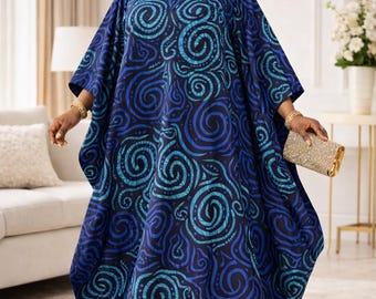 May include: A woman wearing a blue patterned dress and matching head wrap. The dress features a swirling design in shades of blue and teal. She is also wearing gold jewelry and carrying a gold clutch.