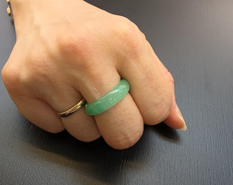 Könnte beinhalten: Ein hellgrüner Jade-Ring wird an einem Finger getragen, neben einem goldenen Band. Der Jade-Ring hat eine glatte, polierte Oberfläche und eine abgerundete Form. Der Ring hat einen lebendigen Grünton.