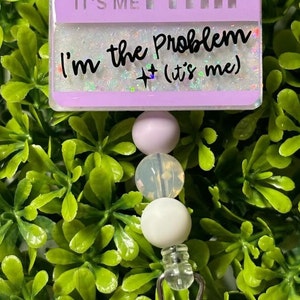 May include: A purple badge holder with the text "It's me Hi... I'm the Problem + (it's me)" with glitter accents. The badge holder is attached to a white and purple beaded lanyard with a silver clip.