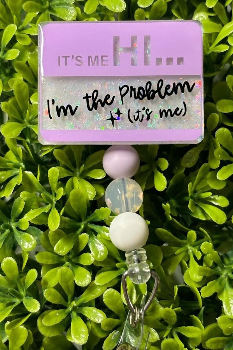 May include: A purple badge holder with the text "It's me Hi... I'm the Problem + (it's me)" with glitter accents. The badge holder is attached to a white and purple beaded lanyard with a silver clip.
