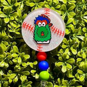 May include: A white baseball-themed badge reel with a green, red, and blue fuzzy monster character. The reel has red stitching and a white background with glitter. The badge reel has three beads: red, blue, and green.
