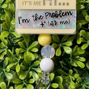 May include: A yellow, white, and clear beaded retractable badge holder with a yellow and white rectangular tag that reads "It's me Hi... I'm the Problem + (it's me)"