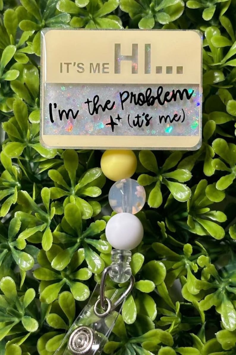 May include: A yellow, white, and clear beaded retractable badge holder with a yellow and white rectangular tag that reads "It's me Hi... I'm the Problem + (it's me)"