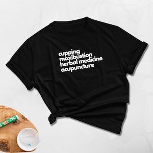 May include: Black t-shirt with white text listing "cupping, moxibustion, herbal medicine, acupuncture." The shirt is folded and knotted at the bottom. A wooden surface with acupuncture needles and a green tube is in the background.