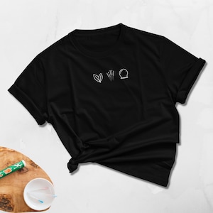 May include: Black t-shirt with a white minimalist design featuring a leaf, needles, and a sphere. The shirt is folded and knotted at the bottom. The background is a light, textured surface.