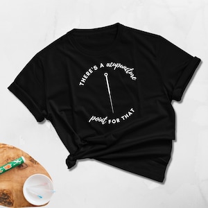 Könnte beinhalten: Schwarzes T-Shirt mit dem Text "THERE'S A acupuncture point FOR THAT" in einem Kreis, zusammen mit einer stilisierten Akupunkturnadel. Das T-Shirt ist gefaltet und am unteren Ende geknotet.