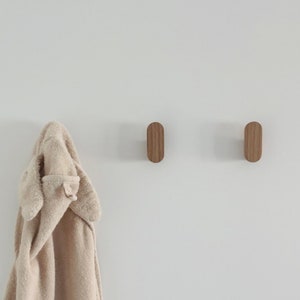 Peut inclure: Deux patères en bois de couleur marron clair sont fixées sur un mur blanc. Une serviette à capuche beige est suspendue à l'un des crochets. Les crochets ont un design simple et minimaliste.