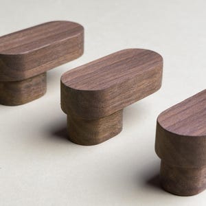 以下が含まれることがあります： 薄い茶色の木製のキャビネットノブが3つあります。ノブは楕円形で、丸みを帯びたトップと正方形のベースがあります。