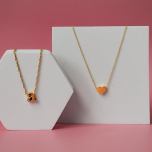 May include: Two gold necklaces with orange pendants are displayed on white geometric pedestals against a pink background. One necklace features the letter "a", and the other has a heart-shaped pendant. The necklaces are delicate and stylish.