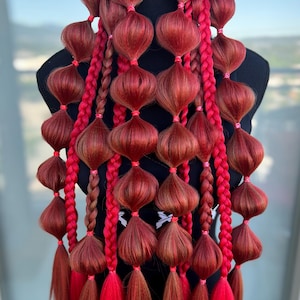 May include: A hair accessory featuring multiple strands of auburn hair styled into bubble ponytails and vibrant red braided sections. The hair is secured with small pink elastic bands at regular intervals, creating a series of rounded segments.