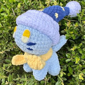 May include: A light blue crocheted plush toy featuring a soft texture, wearing a dark blue nightcap with yellow star accents and a white pom-pom. The figure has yellow oval eyes, a yellow scarf around its neck, and dark blue embroidered facial details.