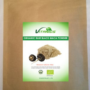 Op de afbeelding: Een bruine papieren zak met een groen en wit etiket met de tekst "VITANICS ORGANIC RAW BLACK MACA POWDER". Het etiket toont ook een hoop bruin poeder en twee zwarte maca-wortels. Product herkomst: Peru.