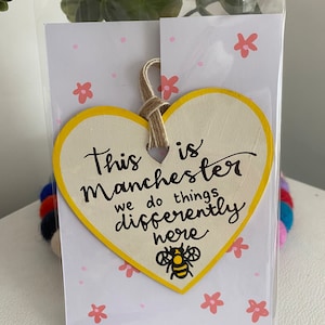 May include: A yellow heart-shaped wooden sign with the text "This is Manchester we do things differently here" and a black and yellow bee illustration. The sign is hanging from a string and is displayed on a white background with pink flowers.