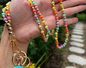 May include: A colorful beaded lanyard with a gold chain and a rainbow-colored tassel keychain. The lanyard is made of pastel-colored beads and gold beads.