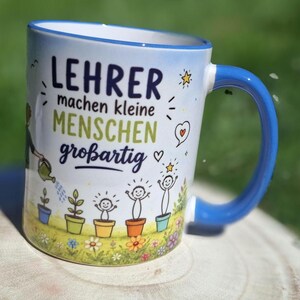 May include: White ceramic mug with a blue handle and rim, featuring the German text "LEHRER machen kleine MENSCHEN gro&szlig;artig" in blue and green. The mug has a design of cartoon plants and people, with a watering can and stars.