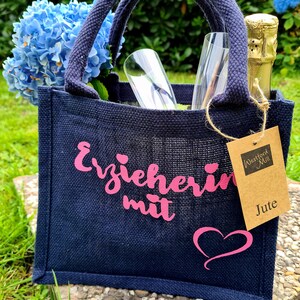 Könnte beinhalten: Eine marineblaue Jute-Tragetasche mit rosa Schriftzug "Erzieherin mit" und einem Herz. Die Tasche enthält zwei Sektgläser, eine Champagnerflasche und blaue Blumen. Ein Etikett lautet "Westford Mill Jute."