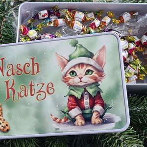 Könnte beinhalten: Eine weiße rechteckige Dose mit Deckel, auf dem eine Cartoon-Katze in einem Elfenkostüm abgebildet ist. Die Dose ist mit verpackten Süßigkeiten gefüllt. Der Text "Nasch Katze" steht in Rot auf dem Deckel. Ein Stapel Kekse ist ebenfalls abgebildet.