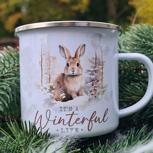 May include: White enamel mug with a silver rim and a white handle. The mug features a watercolor illustration of a brown rabbit in a snowy forest scene. The text "It's a Winterful Life" is written on the mug.