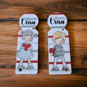 May include: Two white ceramic doorstops with a red, white, and gray striped design. Each doorstop features a cartoon illustration of a child holding a red heart. The top of each doorstop has a circle with the text "Beste Oma" in black.