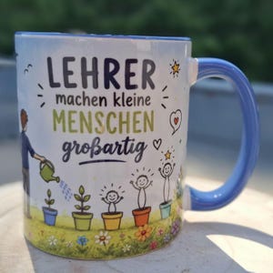 May include: White ceramic mug with a blue handle and rim, featuring the German text "LEHRER machen kleine MENSCHEN gro&szlig;artig" in black and green. The mug has a design of a person watering plants and smiling figures in pots.