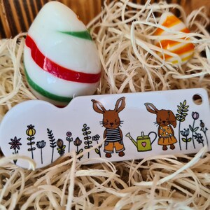 May include: White ceramic key chain with a red and green striped Easter egg, two brown bunnies with flowers and a green watering can, and the text "Happy Easter".