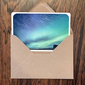 Puede incluir: Una fotografía de paisaje de un cielo nocturno con vetas de luz y una aurora boreal verde y azul se muestra dentro de un sobre marrón. El sobre está sobre una superficie de madera oscura.