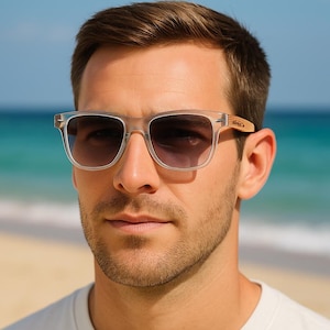 May include: Clear-framed sunglasses with dark lenses. The sunglasses have a rectangular shape and wooden arms. The background shows a beach and ocean. The person in the image is wearing a white t-shirt.