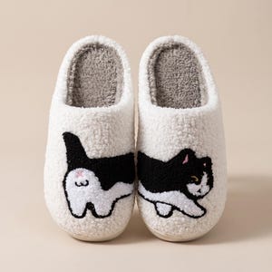 May include: A pair of white fuzzy slippers featuring a black and white cat design. The left slipper shows the back of a cat with a small pink detail, while the right slipper displays the front of a cat. The interior is lined with gray fabric.