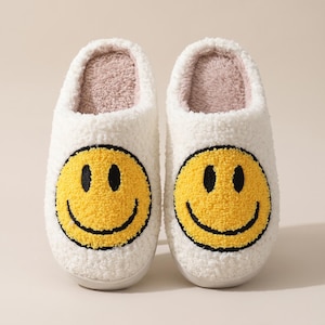 May include: A pair of white fuzzy slippers featuring a large yellow smiley face on the top of each shoe. The smiley face has black embroidered eyes and a black curved mouth. The interior of the slippers is lined with a light beige textured fabric.