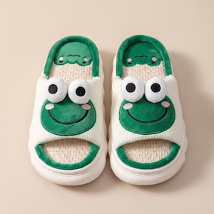 May include: A pair of open-toe slippers featuring a green frog face design with large white eyes, a black stitched smile, and pink cheeks. The slippers have a white ribbed fabric upper, a textured beige footbed, and a thick white rubber sole.