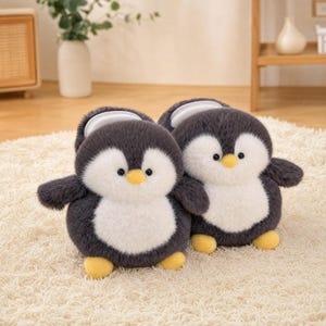 Puede incluir: Un par de pantuflas de peluche con forma de pingüino, confeccionadas en tela suave de color gris oscuro y blanco, con ojos negros redondos y pequeños picos y pies amarillos. Las pantuflas tienen un diseño redondeado con forro interior blanco.