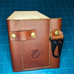 May include: A brown leather pouch with a black metal clip and a small embossed design. The pouch has two brass rivets on the front and a leather strap with two brass rivets.