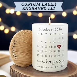 May include: A white candle with a calendar design for October 2026, featuring a highlighted date. The candle has the text "nick & jess" and "the day we said 'I do'". A wooden lid with "LOVE YOU!" is next to the candle.