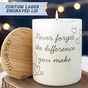 May include: A white candle with the text "Never forget the difference you make" and a stethoscope design. The candle has a wooden lid with "THANK YOU!" engraved. The lid is next to the candle on a wooden surface.