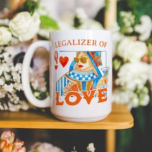 May include: White ceramic mug with a large handle, featuring a colorful illustration of a queen wearing sunglasses and holding a cocktail. The text "LEGALIZER OF LOVE" is printed on the mug. The design includes playing card symbols.