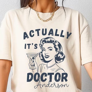 May include: Ivory-colored t-shirt featuring a retro illustration of a woman holding a martini glass. The text reads "ACTUALLY IT'S DOCTOR Anderson" in navy blue. The shirt has a classic, vintage aesthetic.