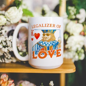 May include: A white ceramic mug featuring a graphic of a king playing card character wearing sunglasses and holding a wine glass. The text reads "LEGALIZER OF LOVE" in orange letters, accompanied by a red heart symbol and the letter K.