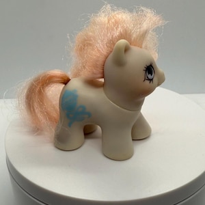 May include: A vintage toy pony figurine in a light cream color with a peach-colored mane and tail. The pony has a blue floral design on its side and detailed eye markings. The toy is standing on a white surface.