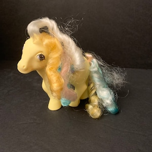 May include: A vintage yellow toy pony with a white and pastel-colored mane and tail. The pony has brown eyes and is posed in a standing position. The hair is curly and styled, with a mix of colors including yellow, pink, and blue.