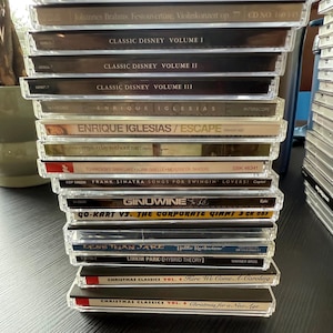 May include: A stack of compact discs in clear plastic cases. Titles include "Classic Disney," "Enrique Iglesias," "Frank Sinatra," and "Linkin Park." The CDs are stacked on a dark surface.