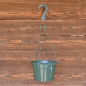 May include: A green hanging plant pot with a decorative swirl pattern. The pot is suspended by a green plastic hanger with a hook. The background is a wooden wall.