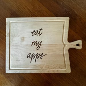 Peut inclure: Planche à découper en bois clair avec une poignée. La planche est de forme rectangulaire avec une rainure sculptée sur le bord. Les mots "eat my apps" sont écrits en cursive.