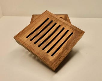 May include: A square wooden soap dish featuring a series of parallel, evenly spaced vertical slats cut into the top surface. The natural light brown wood grain is visible across the flat, elevated platform, which is designed with a sloped edge.
